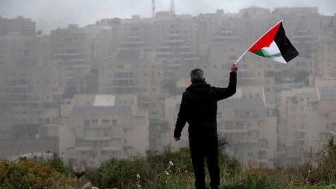 Un palestino agita una bandera frente a un asentamiento judío en Judea y Samaria. (Reuters) Un palestino agita una bandera frente a un asentamiento judío en Judea y Samaria.